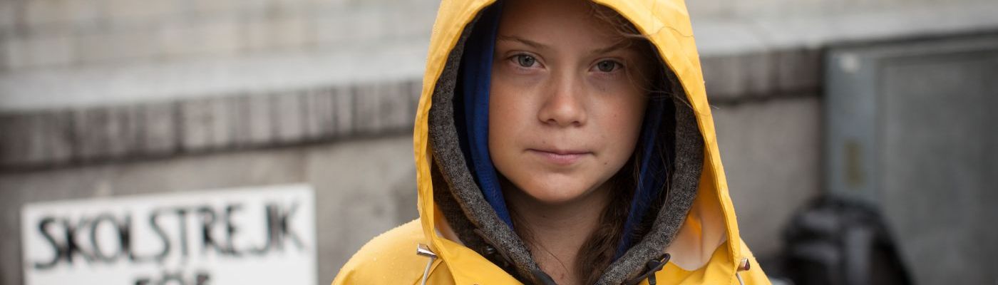 Greta Thunberg steht in einem gelben Regenmantel vor ihrem Protestschild.