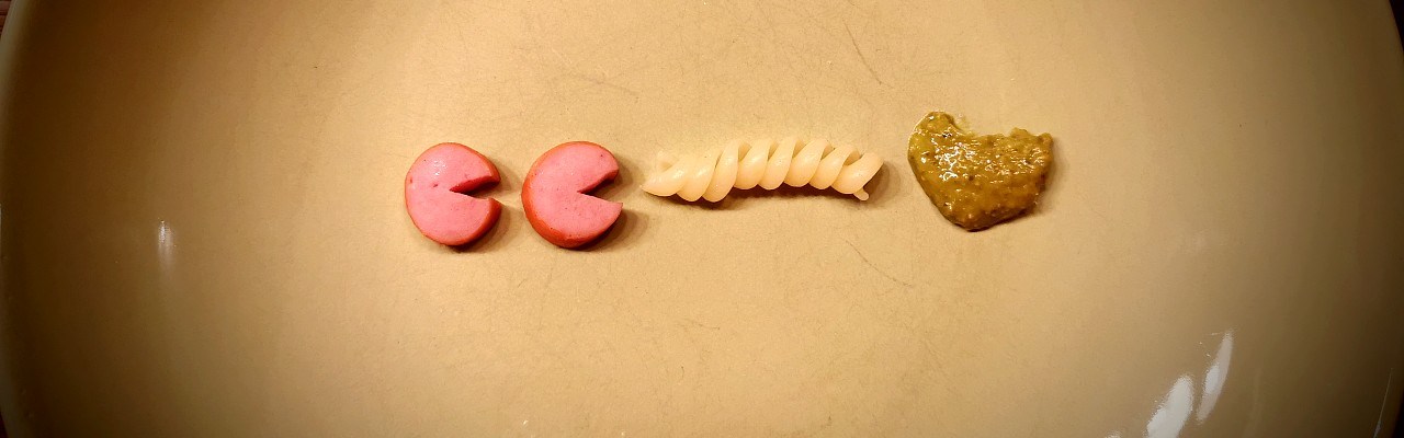 Zwei Stücke Würstchen, eine Nudel und ein Klecks Pesto auf einem grünen Teller.