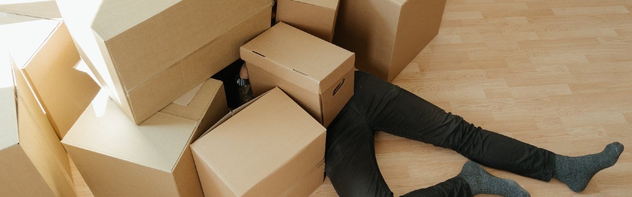 Eine Person in schwarzer Hose liegt unter einem Haufen Pappkartons.
